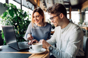 A couple sitting at cafe laughing cheerfully, looking at laptop screen, spending time with pleasure, drinking coffee.