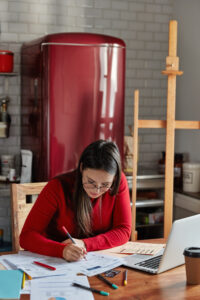 Vertical indoor shot of female writing reports, poses at cozy kitchen with fridge in background.
